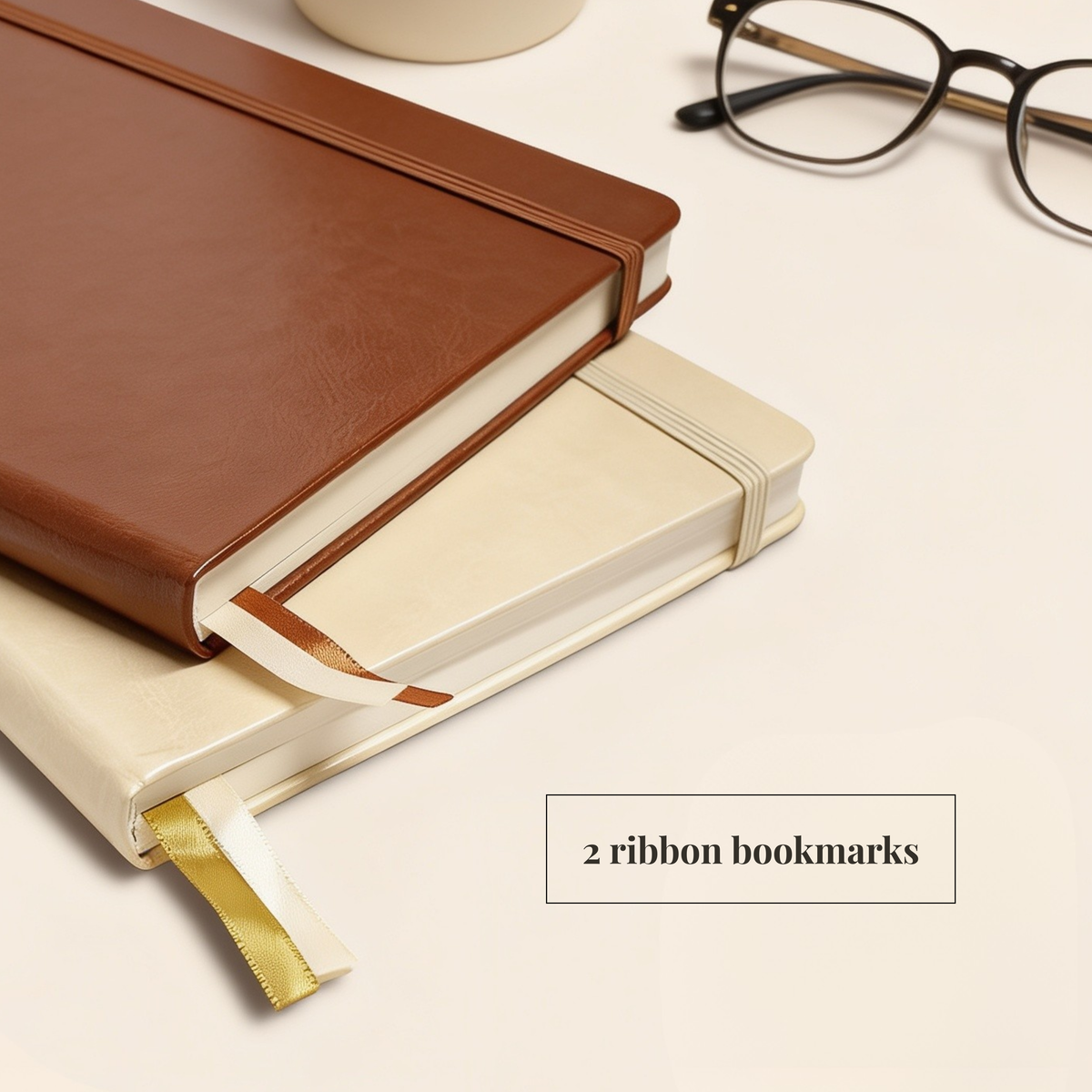 Two hardcover notebooks stacked on a light beige surface, with a brown leather-look notebook on top and a cream notebook underneath. A gold ribbon bookmark is visible from the bottom notebook, and a small text box reads '2 ribbon bookmarks.