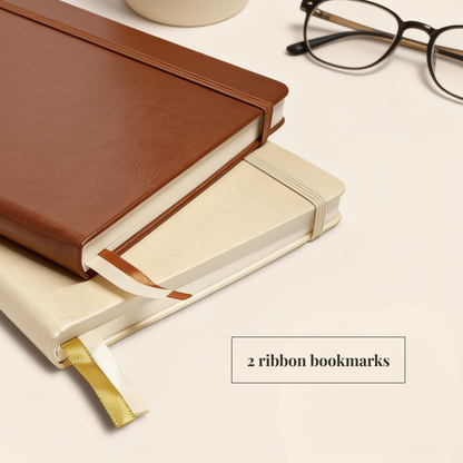 Two hardcover notebooks stacked on a light beige surface, with a brown leather-look notebook on top and a cream notebook underneath. A gold ribbon bookmark is visible from the bottom notebook, and a small text box reads '2 ribbon bookmarks.